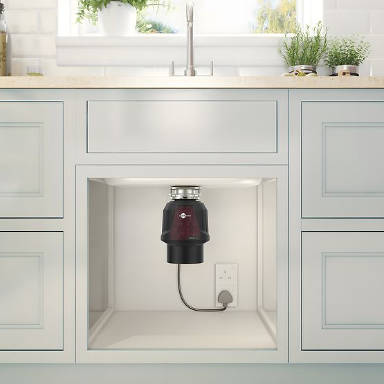 Under-cabinet disposal unit in a kitchen setting with light gray cabinets and a window in the background.